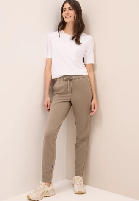 Frau trägt weißes T-Shirt, beige Hose und Sneaker, lehnt entspannt an der Wand.